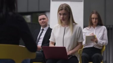 Concentrated secretary typing on laptop keyboard sitting with colleagues as spokesperson talking. Focused beautiful blond Caucasian woman recording meeting in slow motion. Business style concept.