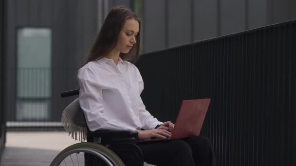 Brunette disabled confident woman messaging online on laptop looking at camera and smiling. Portrait of charming paralyzed Caucasian manager in wheelchair posing outdoors in slow motion. — ストック動画