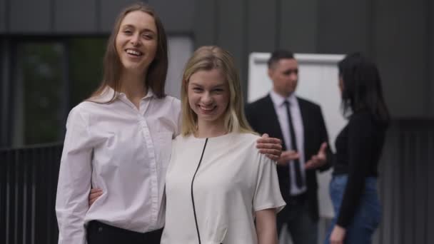 Happy young slim beautiful women looking at camera smiling hugging standing outdoors with colleagues talking at background. Positive Caucasian coworkers posing on meeting. Slow motion. — Vídeo de Stock