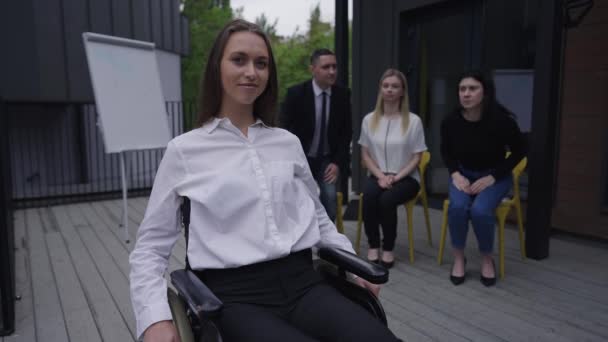 Beautiful confident disabled woman in wheelchair crossing hands looking at camera as colleagues standing up clapping at background. Portrait of satisfied successful Caucasian paraplegic employee. — Video