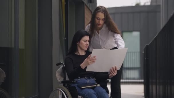 Disabled Caucasian CEO in wheelchair discussing business project with assistant outdoors. Serious successful women talking analyzing strategy. Slow motion. — Vídeo de Stock