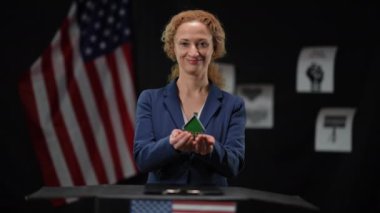 Gülümseyen güzel politikacı kadın oyuncak evi geriyor ve kameraya bakıyor. Kendine güvenen bir bayanın portresi. Siyah arka planda Amerikan bayrağı taşıyan bir emlak programı..