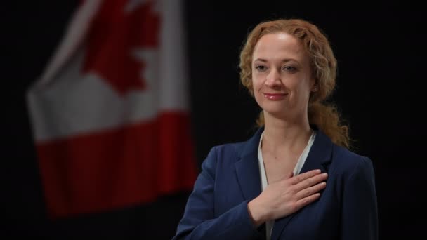 Жінка-політик, тримаючись за груди, слухає державний гімн. Посміхнена жінка стоїть на чорному тлі з канадським прапором. Патріотизм і гордість. — стокове відео