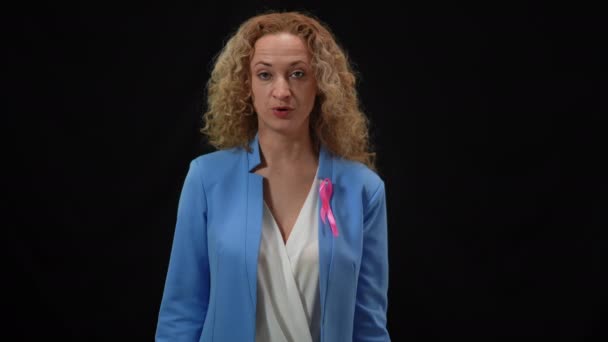 Mulher séria caucasiana falando levantando cartaz consciência do câncer olhando para a câmera. Retrato de orador público explicando perigo de doença em fundo preto. Oncologia e ativismo. — Vídeo de Stock