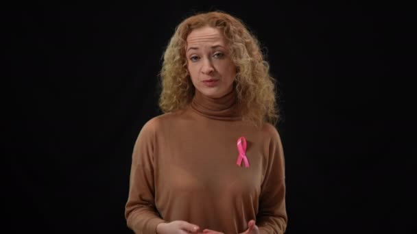 Göğüs kafesinde kanser kurdelesi olan beyaz bir kadının kameraya bakarken orta boy portresi. Hastalık tehlikesini ve korunma önemini açıklayan ciddi bir eylemci. Tıbbi konsept. — Stok video
