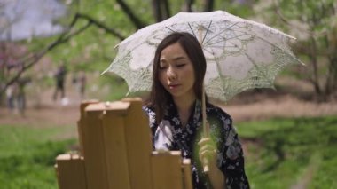 Yakışıklı, gülümseyen Asyalı ressam. Baharda güneşli parkta resim yaparken gülümsüyor. Kimonolu güzel bir Japon kadının portresi. Elinde beyaz güneş şemsiyesi, açık havada ağır çekimde eğleniyor..