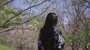 Mutlu, ince, güzel, Asyalı bir kadının bahar parkında yürürken kameraya gülümsemesi. Kendine güvenen mutlu, rahat bir milenyumun portresi yaz günü açık havada poz veriyor. Yavaş çekim.