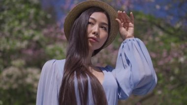 Mavi bluzlu kendine güvenen Asyalı kadın hasır şapkaya dokunuyor güneşli bahar gününde açık havada, lanet olası Sakura parkında kameraya bakıyor. Bahçede poz veren zarif bir Japon hanımın portresi..