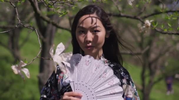 Élő kamera közeledik a magabiztos ázsiai gyönyörű nő gazdaság ventilátor áll a virágzó tavaszi park szabadban. Gyönyörű karcsú hölgy pózol a napsütéses napon, virágzó fákkal körülvéve. Lassú mozgás.. — Stock videók
