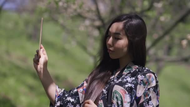 Kimonolu kendine güvenen Asyalı genç bir kadın aynadaki yansımaya bakıyor. Japon milenyumunun bahar aylarında Sakura bahçesinde gün ışığında çekilmiş orta ölçekli görüntüleri.. — Stok video