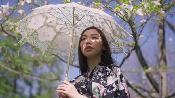 Mittlere Aufnahme einer hinreißenden jungen schlanken Asiatin mit weißem Sonnenschirm im Kimono, die an einem sonnigen Frühlingstag im Park steht und in Zeitlupe Haare berührt. Natürliches Schönheitskonzept. — Stockvideo