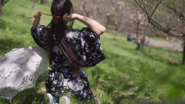 Baksiden av den slanke, vakre asiatiske kvinnen som lager hestehale bundet hår med sløyfe sittende på grønn eng i solrik vårpark. Slank, selvsikker japansk dame i Kimono som hviler utendørs. Langsom bevegelse. – stockvideo