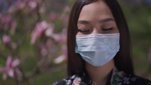 Närbild porträtt av asiatisk ung kvinna i coronavirus ansikte mask öppna ögon tittar på kameran stående i vår park utomhus. Japansk dam poserar i blommande trädgård på Covid-19 pandemi. — Stockvideo