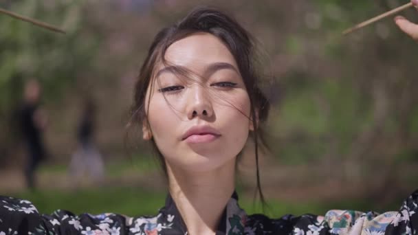 Vista frontal de la cabeza de confianza hermosa mujer joven asiática desatar el pelo en cámara lenta mirando a la cámara. Preciosa dama japonesa atractiva en kimono posando a la luz del sol en el parque de primavera al aire libre. — Vídeos de Stock