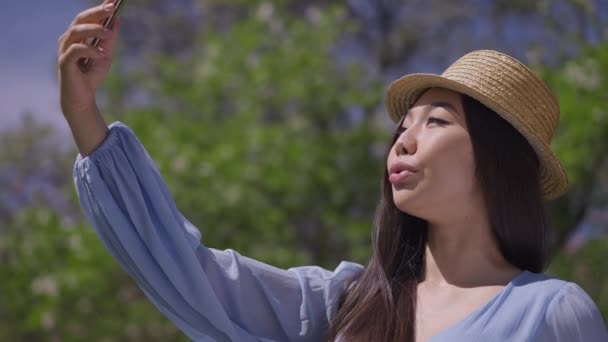 해피 아시아인 여성 이 봄철 공원의 햇살 아래서 스마트폰으로 이야기하고 있습니다. 휴대 전화로 비디오 채팅 앱을 사용하여 편안하고우아 한 천 년의 지푸라기 모자의 모습. 관광 과 생활 방식. — 비디오