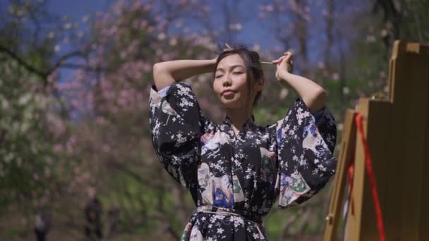 Retrato plano medio de una joven japonesa creativa en kimono haciendo cola de caballo mirando caballete en el floreciente parque de primavera. Confiado sonriendo hermosa esbelta artista pintura al aire libre en la luz del sol. — Vídeos de Stock