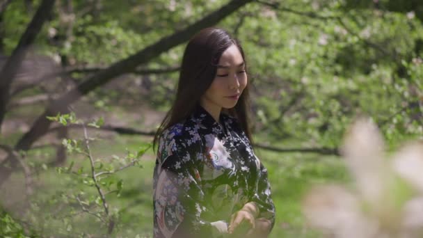 Vakker asiatisk kvinne som står i hagen og ser bort og smiler. Medium shot med nydelig, selvsikker ung dame som nyter solfylt sommerdag utendørs. Skjønnhet og natur konseptet. Langsom bevegelse. – stockvideo