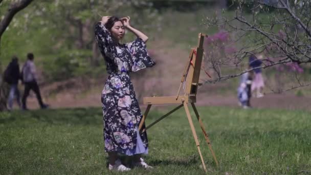 华丽而自信的亚洲女人穿着和服站在春天的公园里，带着画架做辫子。美丽而纤细的日本画家受到户外自然美的启发. — 图库视频影像