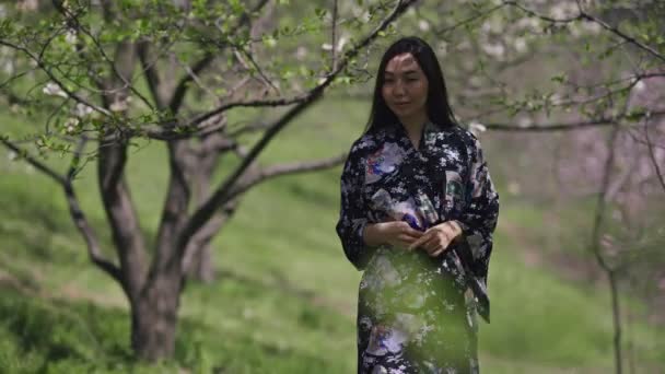 Joven mujer asiática delgada y encantadora en kimono caminando en el soleado parque de primavera girando y sonriendo mirando a la cámara. Retrato de feliz confianza japonesa millennial disfrutando de la belleza de la naturaleza al aire libre. — Vídeo de stock