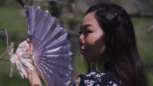 Nahaufnahme von der wunderschönen asiatischen jungen Frau schüttelt Fan im Sonnenlicht im Park wegschauen. Kopfballporträt einer selbstbewussten schönen Dame, die den sonnigen Frühlingstag im Freien lächelnd genießt. — Stockvideo