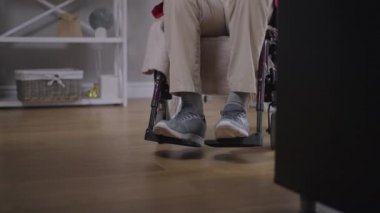 Evdeki oturma odasında ağır çekimde tekerlekli sandalyede oturan sakat kıdemli beyaz adamın bacakları. Kapalı alanda tanınmayan engelli emekliler. Engelli yaşam tarzı kavramı.