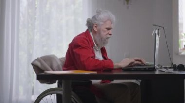 Tekerlekli sandalyede dizüstü bilgisayarda yazan engelli bir adamın zafer jesti yaparken yan görüntüsü. Memnuniyet duyulan özürlü Kafkasyalı emekli kumarhanesi. Yavaş çekim.
