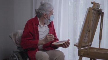 Eski engelli yaratıcı ressam pencereden dışarı bakarak resim yapıyor. Covid-19 yüz maskeli beyaz adamın portresi huzurevinde hobi olarak eğleniyor..