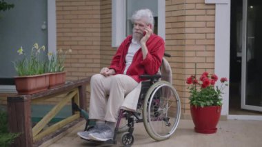 Tekerlekli sandalyedeki mutlu yaşlı adam verandada telefonla konuşuyor. Olumlu, neşeli, Kafkasyalı emekli arkadaşlarıyla ya da aileleriyle akıllı telefondan sohbet ederken geniş açı.