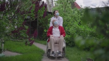 Tıbbi önlüklü ve koronavirüslü genç bir kadın huzurevi bahçesinde engelli bir ihtiyarla tekerlekli sandalye takıyor. Olumlu sempatik Kafkas hemşire yaşlı hasta emeklilerle ilgileniyor..