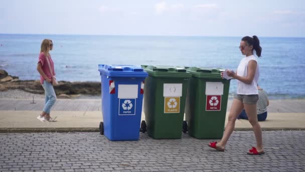 Amplio tiro de mujer joven rasgando papel arrojando basura en un contenedor especial para la separación de la basura en el resort con personas relajadas que pasan al fondo en la costa. Concepto de gestión de la basura y turismo — Vídeo de stock