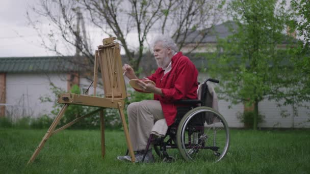 Talentvolle creatieve gehandicapte oude man schilderen op ezel in het voorjaar zomertuin op de achtertuin in slow motion. Side view breed schot van geschoolde geïnspireerde blanke gepensioneerde genieten van hobby buitenshuis. — Stockvideo