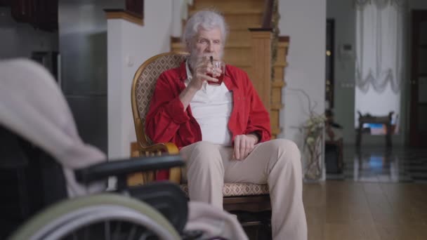 Portret pewnego siebie przystojnego starszego pana pijącego herbatę siedzącego w wygodnym fotelu z rozmazanym wózkiem inwalidzkim z przodu. Siwy, brodaty, biały, niepełnosprawny emeryt w domu, w salonie. — Wideo stockowe
