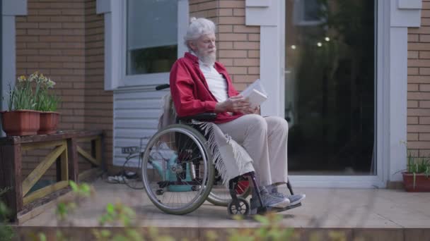 Widok z boku na upośledzonego brodatego seniora czytającego książkę zamykającą i patrzącego w słońce. Szerokie ujęcie portretu inteligentnego białego niepełnosprawnego emeryta na wózku inwalidzkim na podwórku ganek na zewnątrz. — Wideo stockowe