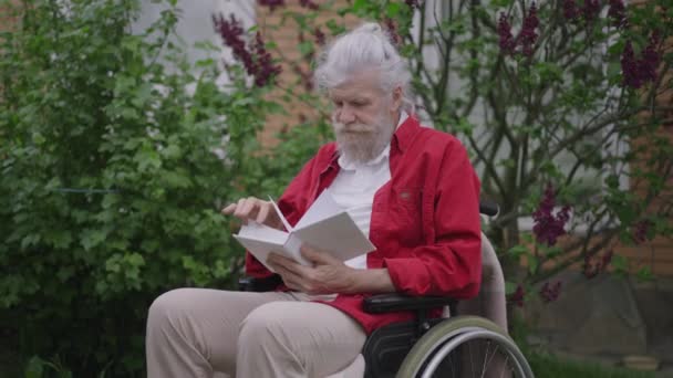 Absorvido velho deficiente lendo livro interessante no jardim do quintal ao ar livre. Retrato de tiro médio de deficiente inteligente aposentado caucasiano desfrutando de hobby ao ar livre em casa. Movimento lento. — Vídeo de Stock