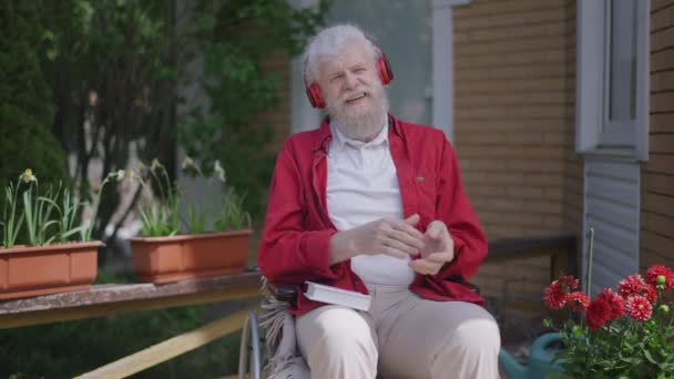 Alegre barbudo discapacitado sénior en silla de ruedas aplaudiendo cantando en cámara lenta escuchando música en auriculares. Retrato de positivo relajado caucásico retirarse disfrutando de hobby en la luz del sol — Vídeos de Stock