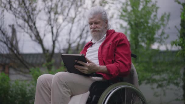 Седовласый бородатый инвалид, старик улыбается, держа дневник. Портрет позитивного пенсионера-инвалида, сидящего в инвалидной коляске на открытом воздухе в весеннем летнем саду на заднем дворе. — стоковое видео