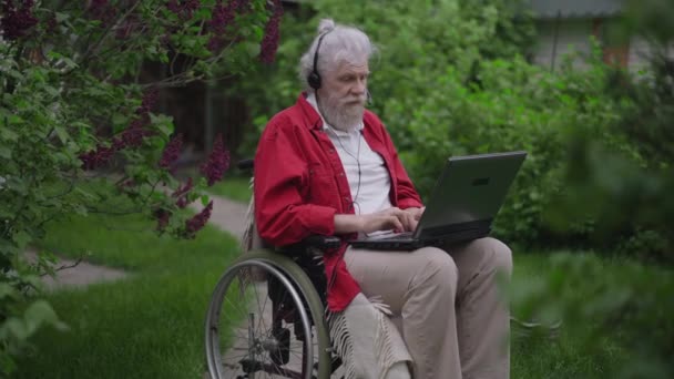 Bonito e alegre homem idoso deficiente em cadeira de rodas sorrindo conversando no bate-papo de vídeo laptop. Retrato de positivo barbudo caucasiano aposentado bate-papo on-line do jardim do quintal. Tecnologias modernas. — Vídeo de Stock
