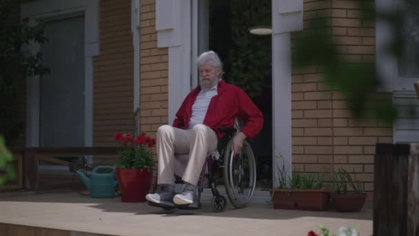 Positiv skäggig handikappad man rulla rullstol på verandan i slow motion beundra solljus utomhus. Brett fotograferat porträtt av glad manlig vit handikappad pensionär på sommaren vårdagen. — Stockvideo