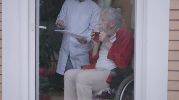 Senior bearded disabled man in wheelchair drinking tea as nurse bringing documents for sign. Portrait of relaxed Caucasian retiree enjoying leisure in nursing home indoors. Slow motion. — 图库视频影像