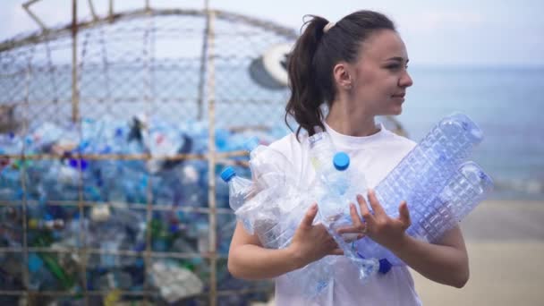 Közepes felvétel fiatal kaukázusi nőről, aki üres műanyag palackokat tart a tengerparton, és körülnéz. Pozitív gyönyörű női önkéntes szemetet gyűjt a tengerparton a szabadban. — Stock videók