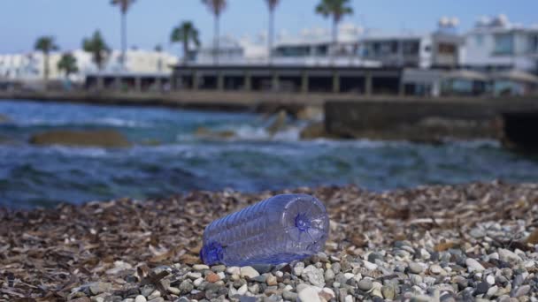 Műanyag üres palack esik lassított felvételen kavicsos strand mediterrán tenger hullámok gördülő tengerparton a háttérben. A környezetszennyezés fogalma a szabadtéri üdülőhelyeken. — Stock videók