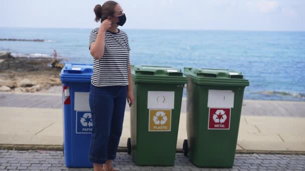 Mujer joven caucásica de confianza que se quita la máscara de cara coronavirus tirar basura en cubo de basura de plástico en la costa del mar y alejarse en cámara lenta. Millennial separando la basura en el complejo turístico. — Vídeo de stock