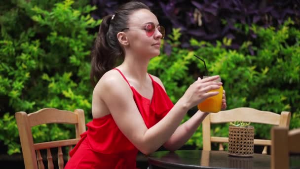 Впевнена, що вродлива жінка - туристка п "є освіжаючий вітаміновий фруктовий коктейль, який сидить на відкритому повітрі в кафе на середземноморському курорті. Світла жінка в червоній сукні відпочиває в сонячний літній день.. — стокове відео