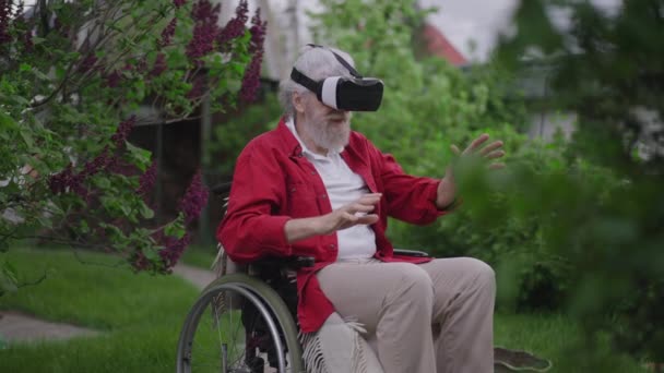 Anciano discapacitado con auriculares VR sentado en silla de ruedas al aire libre moviendo las manos en cámara lenta sonriendo. Positivo feliz discapacitado caucásico jubilado disfrutando de la realidad aumentada en el jardín de primavera en el patio trasero. — Vídeo de stock