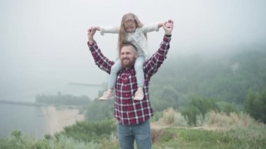 Gülümseyen genç sakallı adam ellerini uzatıyor, kızı omuzlarına alıyor ve başka tarafa bakıyor. Heyecanlı beyaz bir babanın portresi. Küçük tatlı kızıyla gezinin keyfini çıkarıyor..