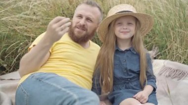 Hasır şapkalı rahat küçük kız sakallı adamla çayırda oturuyor. Tasasız, güzel, beyaz bir kızın hafta sonu dışarıda babasıyla eğlenmesi portresi. Çocukluk eğlencesi..