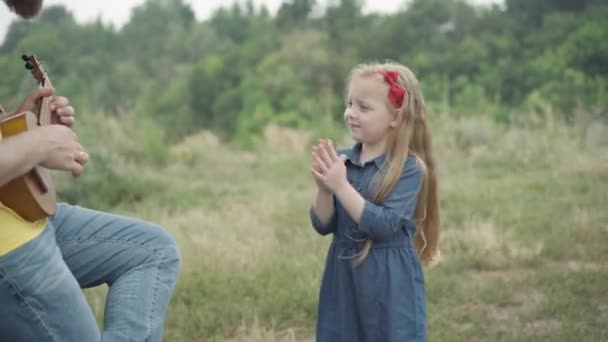 Joyeux petite fille applaudissements soutenant homme méconnaissable jouant ukulélé. Portrait de fille caucasienne mignonne positive approuvant père divertissant enfant à l'extérieur. Concept de joie et de loisirs. — Video