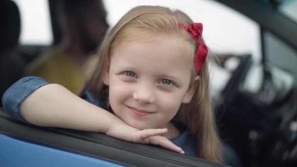 Közelkép egy gyönyörű fehér szőke kislányról, aki autóban ül, és homályos férfival a háttérben a kamerába néz. Fejlövés egy boldog, csinos lányról apával az autóban. Családi fogalom. — Stock videók