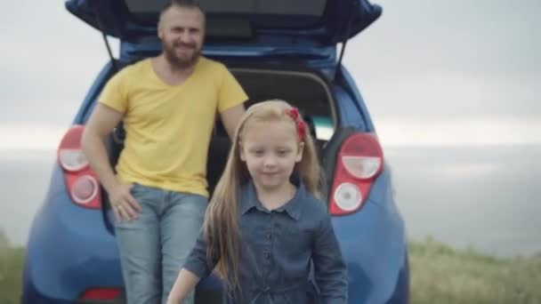 Cute cheerful little girl running along summer meadow on river bank with blurred father in car trunk at background. Positive Caucasian pretty child having fun travelling with parent. Live camera. — Stock Video