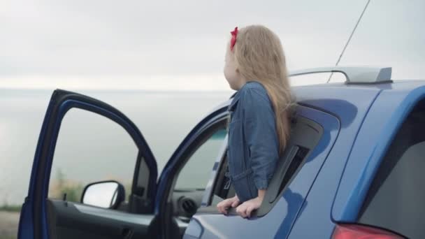 Entusiasmada menina bonita feliz saindo do carro olhando ao redor admirando a beleza da natureza no dia de outono nebuloso. Retrato de criança caucasiana alegre descansando na margem do rio ao ar livre rindo. — Vídeo de Stock